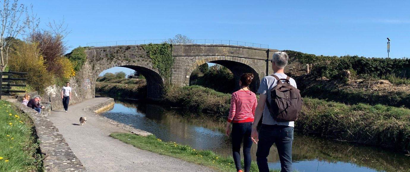 a picture of 2 pilgrims walking along the river. There is a bridge across the river